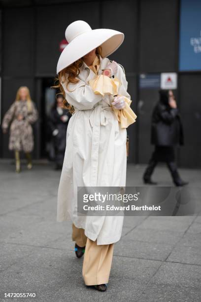 Guest wears a white felt / wool oversized hat, a beige shirt, matching beige suit pants, a white belted long trench coat, a brown and black LV...