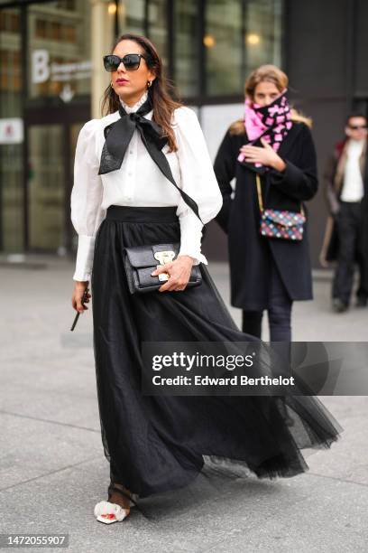 Guest wears black sunglasses, gold earrings, a black silk long knot scarf, a white ruffled high neck blouse, a black high waist tulle long skirt, a...