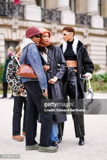 Model wears black sunglasses, a white hoodie sweater, a blue faded denim jacket, a camel shiny varnished leather shoulder bag, black denim pants,...