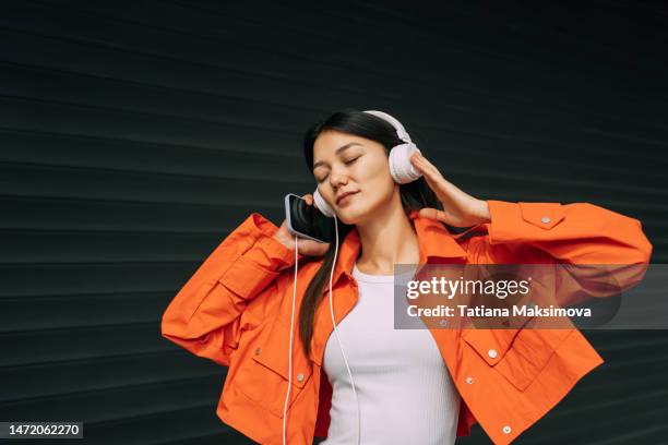 portrait of beautiful asian dancing woman in orange jacket and white headphones on black background. - headphones asian fotografías e imágenes de stock