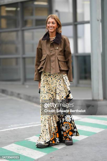 Guest wears gold large chain necklace, a brown denim cargo jacket, a pale yellow and black / white / orange flower print pattern ruffled long skirt,...