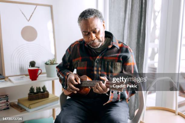 lässig gekleideter älterer mann, der in seiner wohnung ukulele spielt - ukulele stock-fotos und bilder