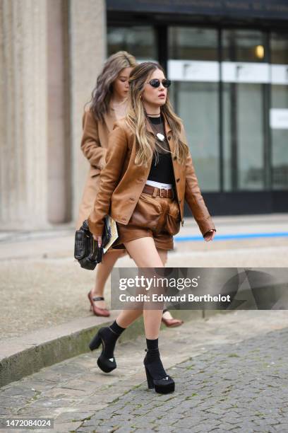 Paola Alberdi wears black sunglasses, a black tulle t-shirt, a black tank-top, a camel shiny leather buttoned jacket, camel shiny leather belted...