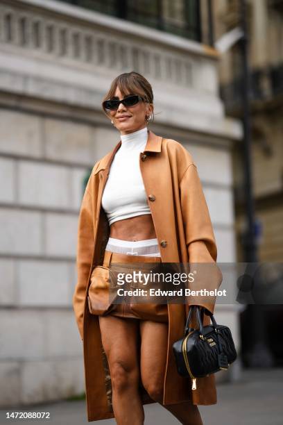 Camila Coelho wears black sunglasses, silver earrings, a white high neck / cropped tank-top from Miu Miu, a camel shiny leather long coat from Miu...