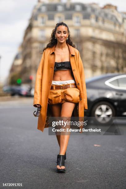 Tamara Kalinic wears brown sunglasses from Miu Miu, a black belted shoulder-off / cropped top from Miu Miu, a camel shiny leather long coat, a camel...
