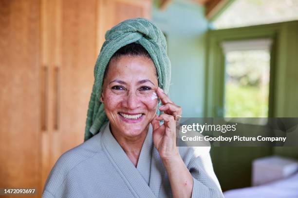 smiling mature woman in a bathrobe and towel applying moisturizer to her cheek - exfoliation stock pictures, royalty-free photos & images