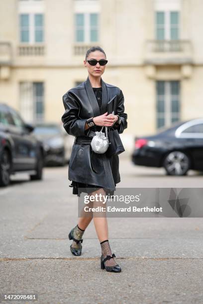 Sophia Roe wears black sunglasses, a black short dress, a black shiny leather belted coat, a black shiny with gold leather 5 embroidered pattern...