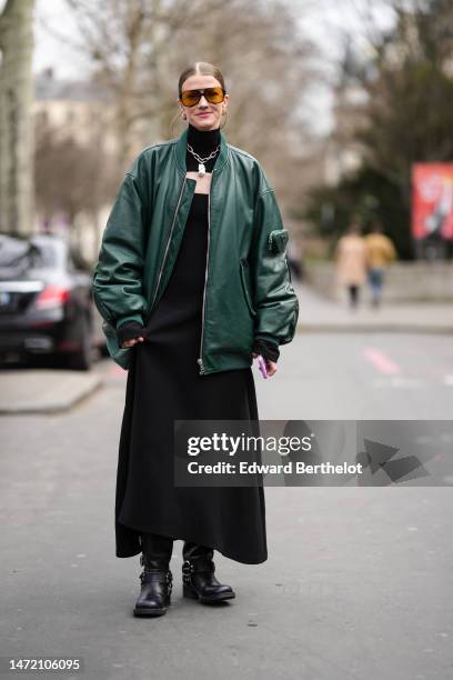 Marianne Theodorsen wears black and orange sunglasses, a black turtleneck / cut-out chest / long dress, a dark green shiny leather oversized bomber...