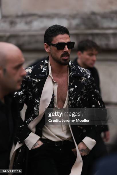 Michele Morrone wears Dolce & Gabbana black sunglasses, a black sequin suit jacket, a beige satin shirt with a scarf detail, black pants and a black...