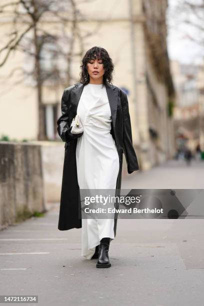 Xiayan wears a white wrap long dress, a black shiny leather long coat, a beige embossed leather puffy handbag, black satin gloves, black shiny...