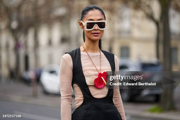 Pornwika Spiecker wears beige large squared sunglasses, a beige long sleeves t-shirt, a red shiny leather large flower pendant necklace, a black...