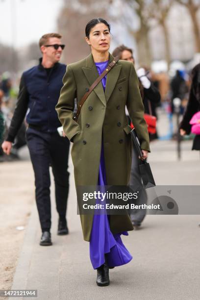 Caroline Issa wears a neon purple long dress, a khaki wool buttoned long coat, a dark brown shiny leather crossbody bag, black shiny leather slit /...