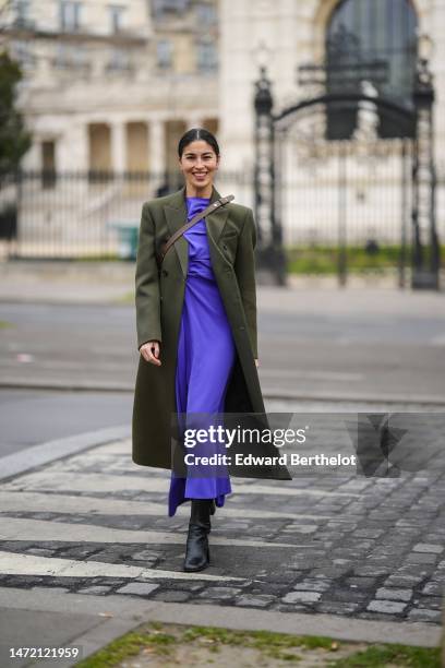 Caroline Issa wears a neon purple long dress, a khaki wool buttoned long coat, a dark brown shiny leather crossbody bag, black shiny leather slit /...