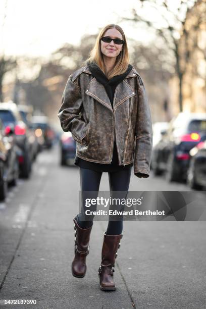 Pernille Teisbaek wears black sunglasses, a black hoodie sweater, a dark brown faded leather oversized jacket, black nylon legging pants, a dark...