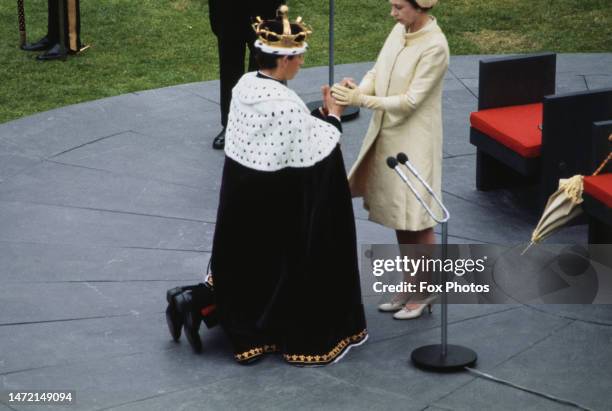 British Royal Elizabeth II places the gold coronet onto the head of her son, Charles, Prince of Wales, who wears a ceremonial robe trimmed with...