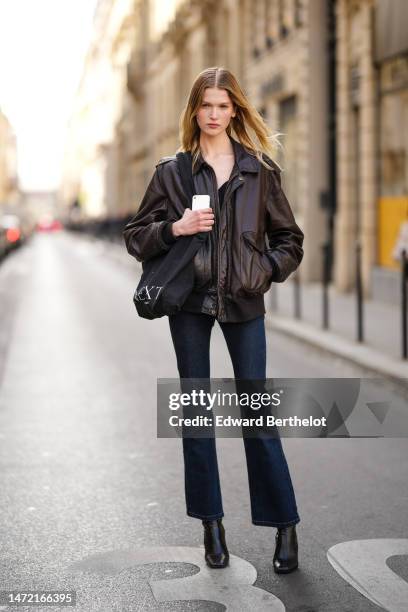 Guest wears a black t-shirt, a dark brown shiny leather zipper bomber coat, a black cotton shoulder bag, navy blue denim flared pants, black shiny...