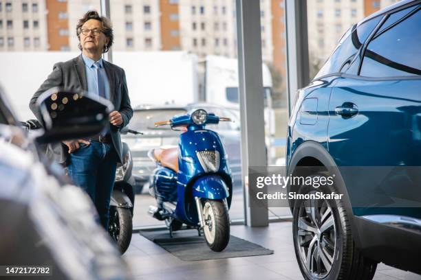 businessman is standing next to a new car at his car dealership,looking at a customer in the distance with serious facial expression,scooters in the back - motorbike shop stock pictures, royalty-free photos & images