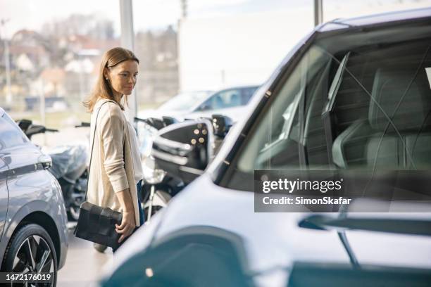 female customer is standing indoors at a car dealer,looking at a new suv,family car behind her,standing at the side of the suv - autodealer stockfoto's en -beelden