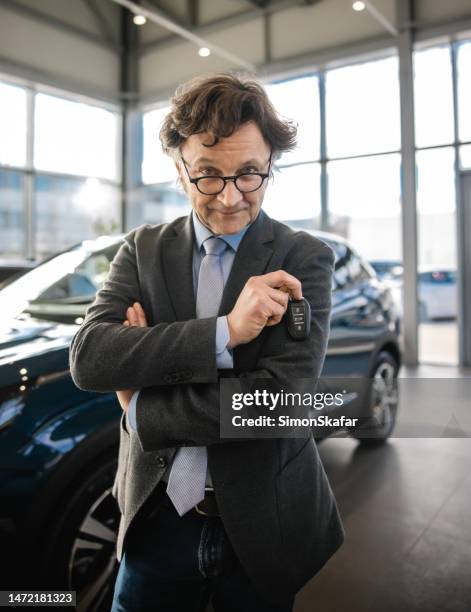 geschäftsmann steht drinnen in seinem autohaus, schaut mit ernstem gesichtsausdruck in die kamera, hält einen autoschlüssel in der hand, suv im hintergrund - car dealer stock-fotos und bilder