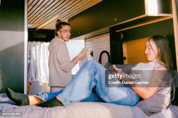 girl using her mobile while her boyfriend while cooking for her in the campervan, couple in love living in a campervan, valentine's day, love in a campervan - cooking demo stock pictures, royalty-free photos & images