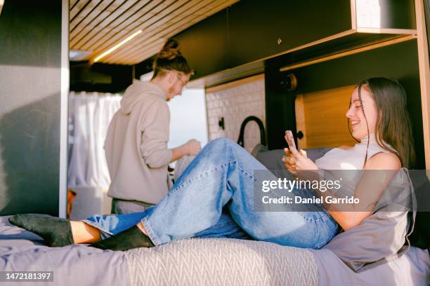 girl using her mobile while her boyfriend while cooking for her in the campervan, couple in love living in a campervan, valentine's day, love in a campervan - cooking demo stock pictures, royalty-free photos & images