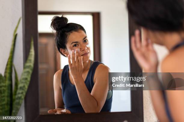 woman applying white sunblock to face, looking at
 bathroom mirror - mature women skincare stock pictures, royalty-free photos & images