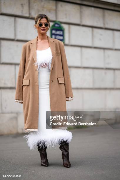 Guest wears black circle sunglasses, a beige long coat from Miu Miu, a white with embroidered feathers V-neck / cropped top, a matching white high...