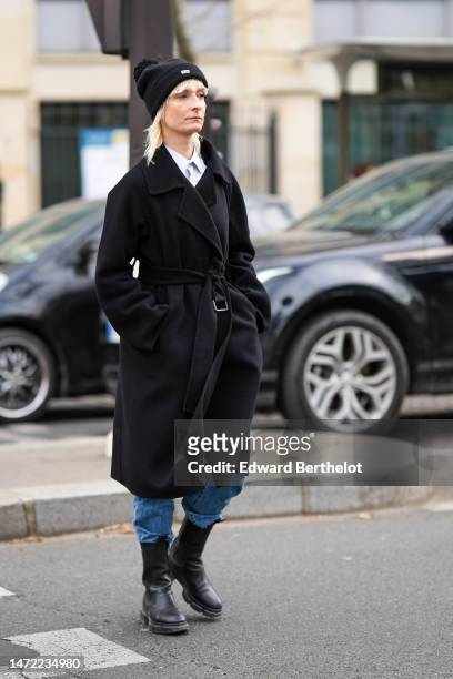 Guest wears a black wool with embroidered pom pom beanie, a white shirt, a white tie, a black wool long belted coat, blue denim ripped pants, black...