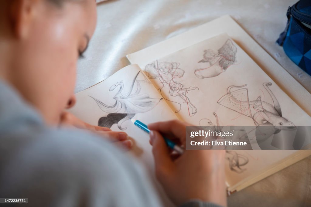 Teenager sitting at a dining table, sketching with a pencil in a notebook.
