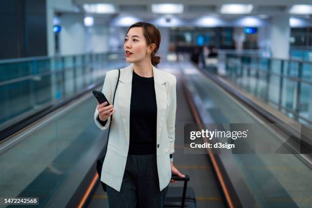 professional young asian businesswoman carrying her suitcase, using smartphone while walking at the airport. business trip. business travel. travel and vacation. business on the go concept - ir hacia delante fotografías e imágenes de stock