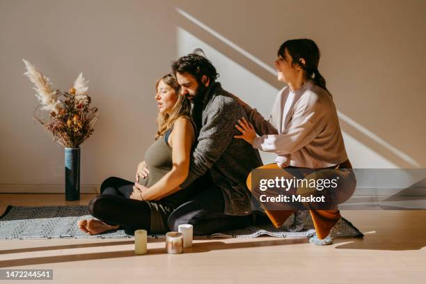 female instructor crouching while teaching yoga to expectant couple in exercise room - pregnancy class stockfoto's en -beelden