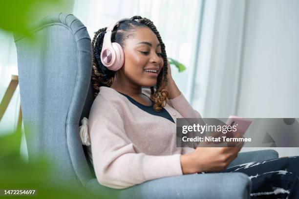 music can improve your mental health in daily life. side view of an african american young woman enjoying listening to a podcast or musing over headphones by using a smartphone during relaxing on a sofa at home. - podcast headphones stock pictures, royalty-free photos & images