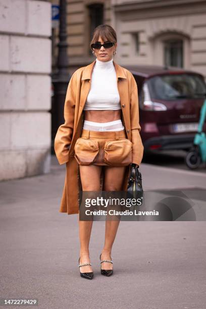 Camila Coelho wears a white turtleneck crop top, tan leather Miu Miu mini skirt, tan leather coat, black bag and shoes, outside Miu Miu, during Paris...