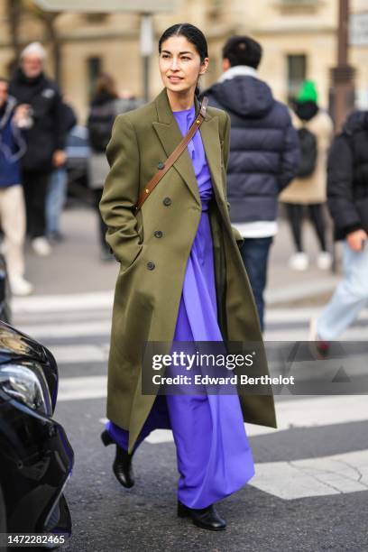 Caroline Issa wears a neon purple long dress, a khaki wool buttoned long coat, a dark brown shiny leather crossbody bag, black shiny leather slit /...