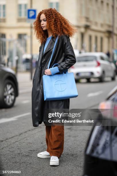 Guest wears a blue sweater, a black shiny leather long coat, camel brown large denim pants, a blue shiny leather embossed logo large shoulder bag...