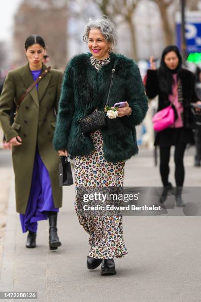 Guest wears gold earrings, a beige with black and multicolored leopard print pattern long dress, a dark green fluffy fur jacket, a black shiny...