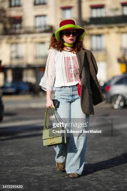 Guest wears a red felt wool hat, a green silk scarf, black sunglasses, a white with embroidered red braided pattern blouse, a brown blazer jacket,...