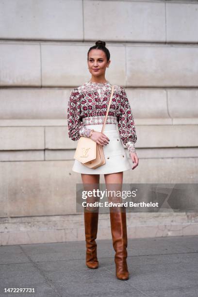 Alicia Vikander wears white pearl and gold earrings, a white with red and black flower print pattern pleated / long puffy sleeves blouse, a white...