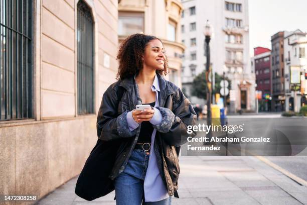 smiley afro woman using a mobile walking in the street - street style foto e immagini stock