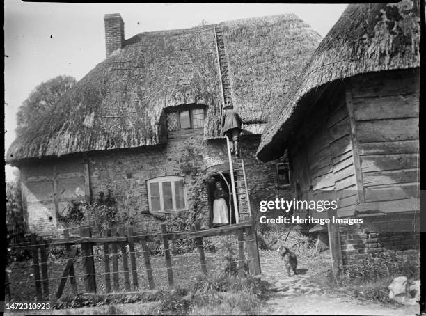 Wootton Rivers, Wiltshire, 1923. A thatcher working on the roof of an unidentified cottage in Wootton Rivers while a woman stands in the doorway. In...