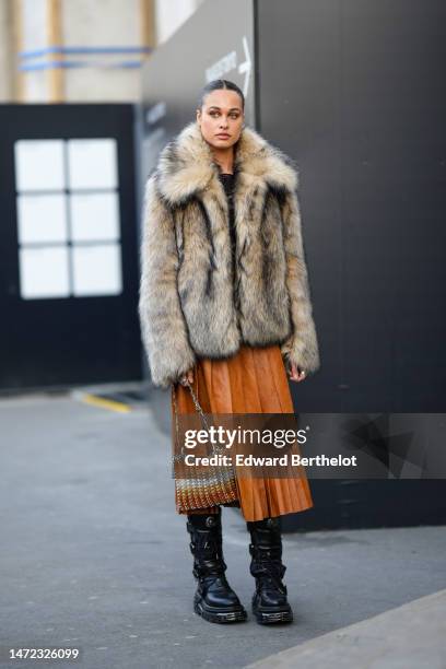 Guest wears gold earrings, a black lace print pattern velvet top, a brown fur coat, a camel shiny leather pleated / accordion midi skirt, a silver...
