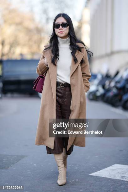Guest wears black sunglasses, a white turtleneck pullover, a beige wool long coat, a burgundy matte leather shoulder bag from Chanel, a black shiny...