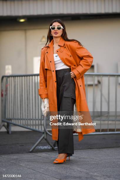 Guest wears white sunglasses, gold earrings, an orange shiny leather long coat, a white tank-top from Prada, a beige braided shiny leather Jodie...