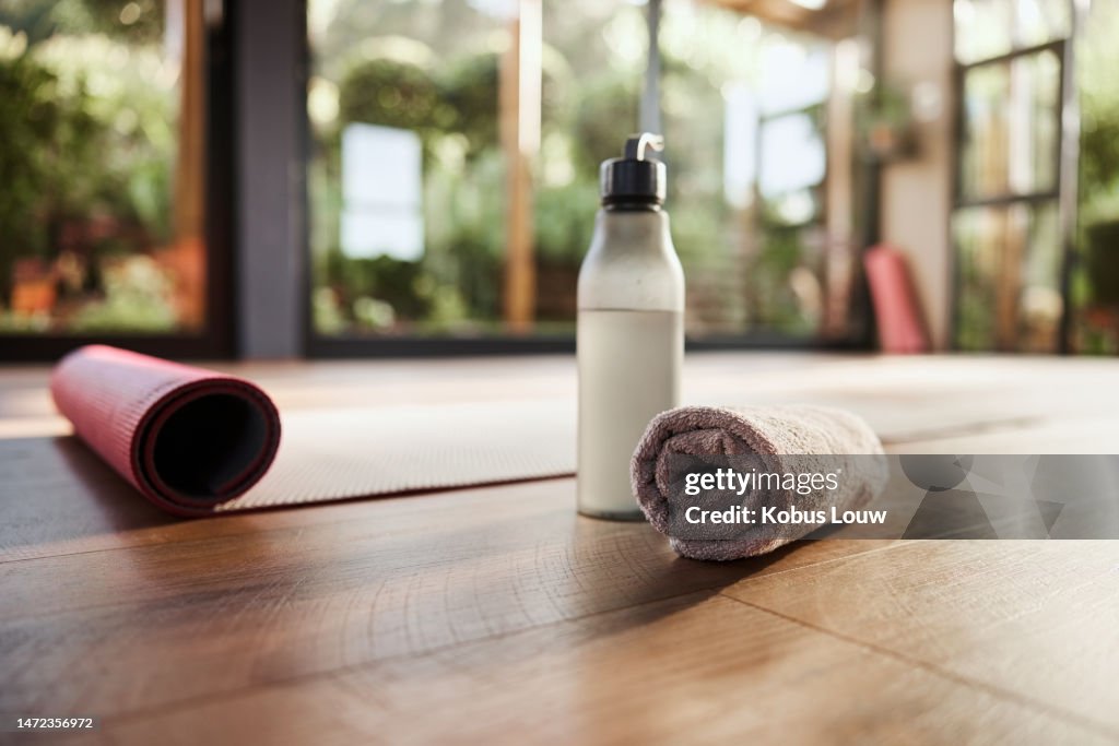 Wasser, Handtuch und Yogamatte im leeren Fitnessstudio für Wellness, psychische Gesundheit oder innere Ruhe hautnah. Fitness, Bewegung und Pilates mit Stilllebengeräten auf dem Boden eines Fitnessstudios für ein Training