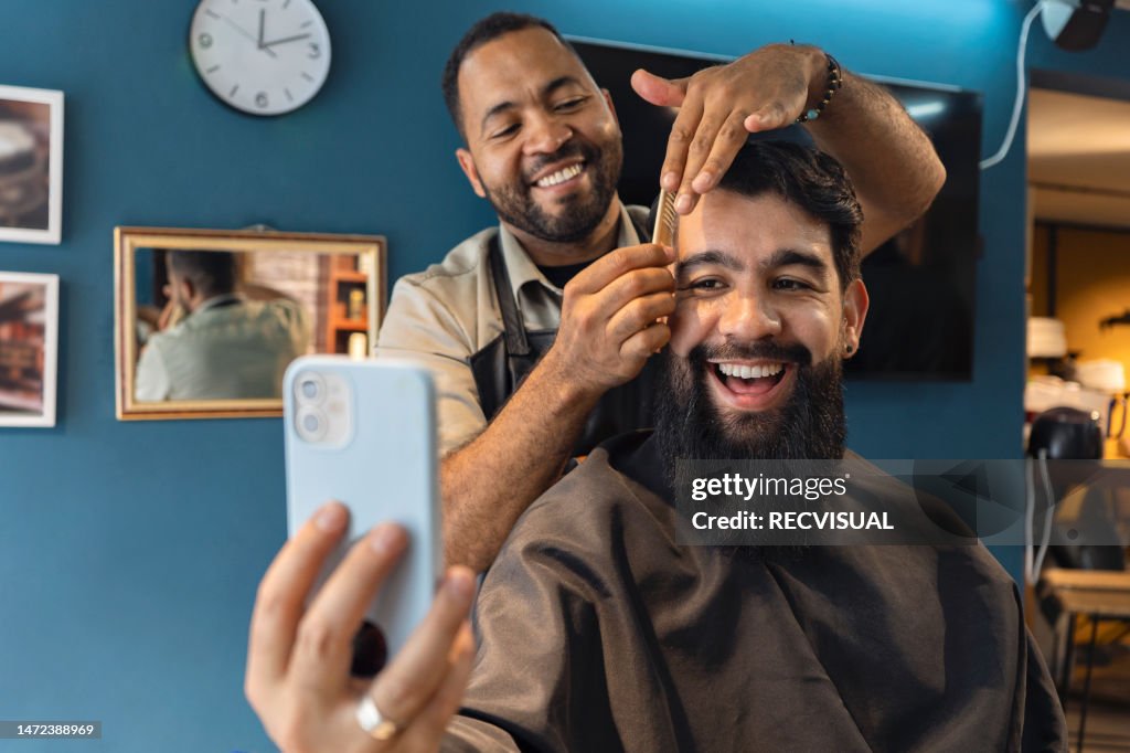 Lächelnder Friseur und Kunde im Friseursalon schaut sich ein Video auf einem Smartphone an.