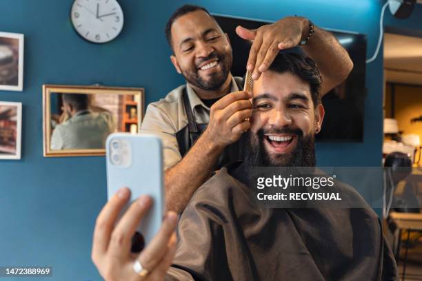 smiling barber and customer in the barbershop watching a video on a smartphone. - barber stock pictures, royalty-free photos & images