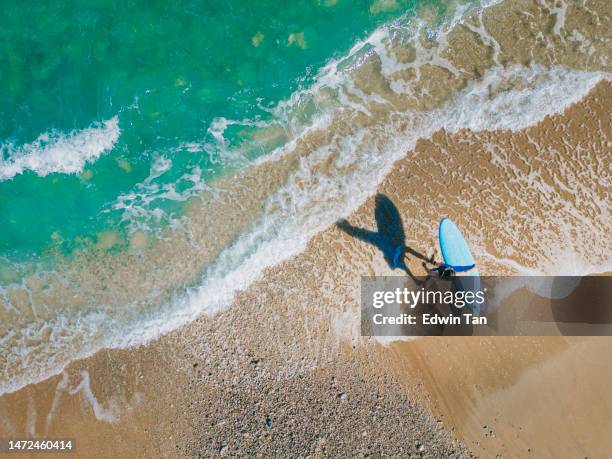 asian chinese male surfer walking into sea preparing for surfing riding wave - beach bird's eye perspective imagens e fotografias de stock