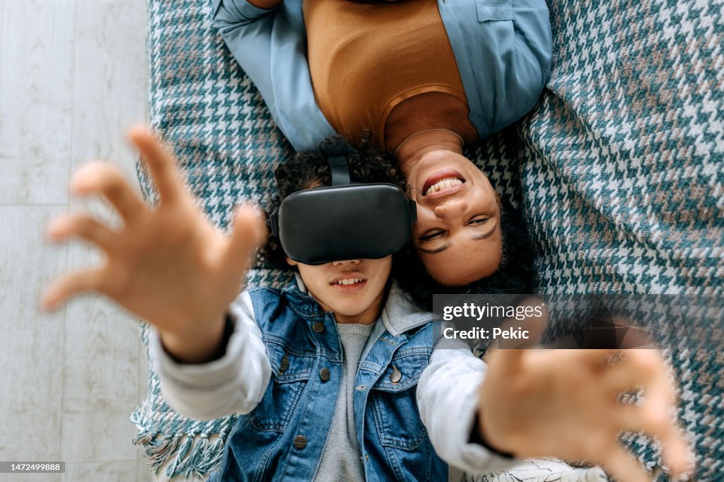 Vista de alto ángulo de un hijo adolescente y una madre usando auriculares de realidad virtual mientras están acostados en un sofá