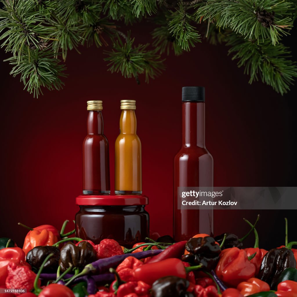 Various types of hot sauce. Bottles and jar of sauce among various types of peppers under spruce branches. Pepper pods. Hot pepper. Spicy food. Red sauce. Chili ketchup. Dark red background. Side view