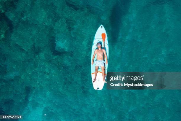 drone view of man lying on a stand up paddle in summer and floating on beautiful sea - aerial beach view sunbathers stock pictures, royalty-free photos & images